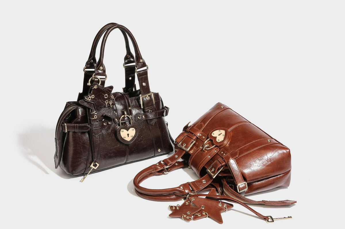 Structured leather-look satchel bag with heart padlock charm, star charm, gold-tone buckle hardware, dual top handles, and