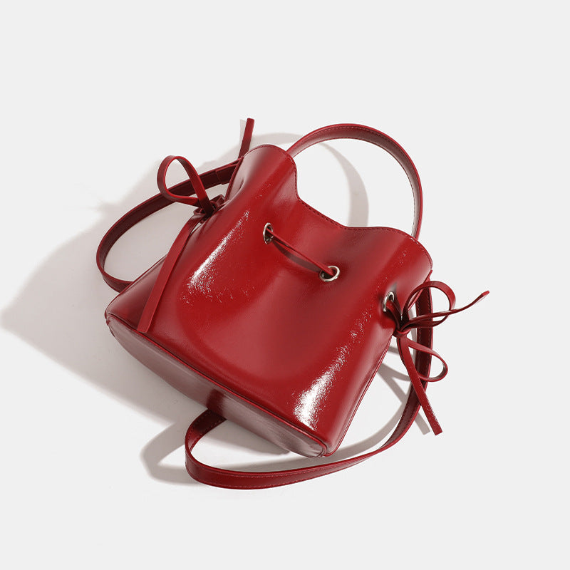 Compact glossy leather-look bucket bag with drawstring bow-tie closure, rounded top handle, and silver-tone grommet hardware