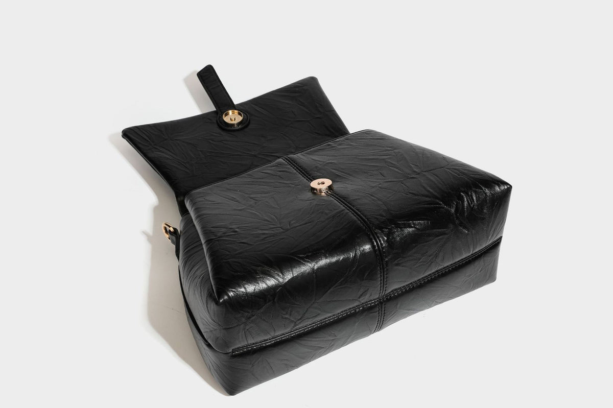 Structured black satchel bag with glossy crinkled leather-look finish, gold-tone emblem flap closure, slim top handle, and