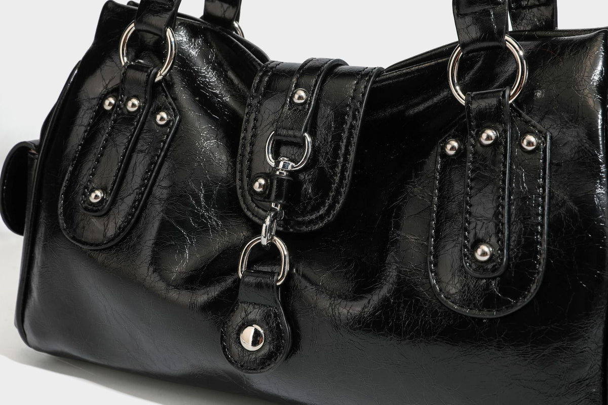 Glänzende, geknautschte Barrel-Satchel mit zwei oberen Tragegriffen, vorderem Klappen-Riemen-Panel, Ring- und Nietenbeschlägen sowie einem abnehmbaren