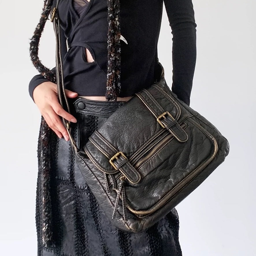 Black distressed leather-look flap crossbody bag with dual buckle straps, front zip pocket, and antique gold-tone hardware