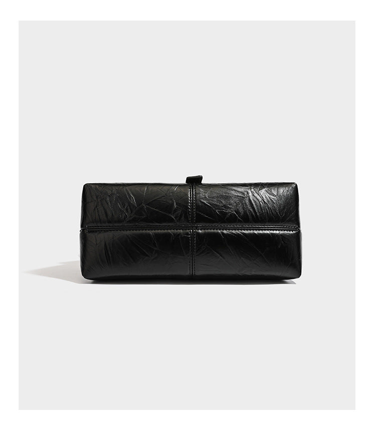 Structured black satchel bag with glossy crinkled leather-look finish, gold-tone emblem flap closure, slim top handle, and