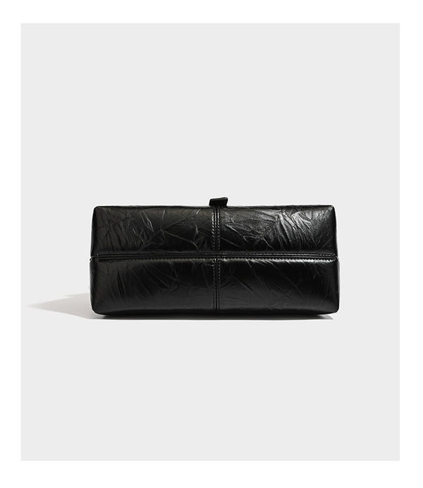 Structured black satchel bag with glossy crinkled leather-look finish, gold-tone emblem flap closure, slim top handle, and