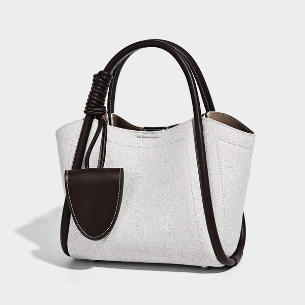 Light blue denim trapezoid tote bag with black leather-look trim, knotted top handles, detachable crossbody strap, front