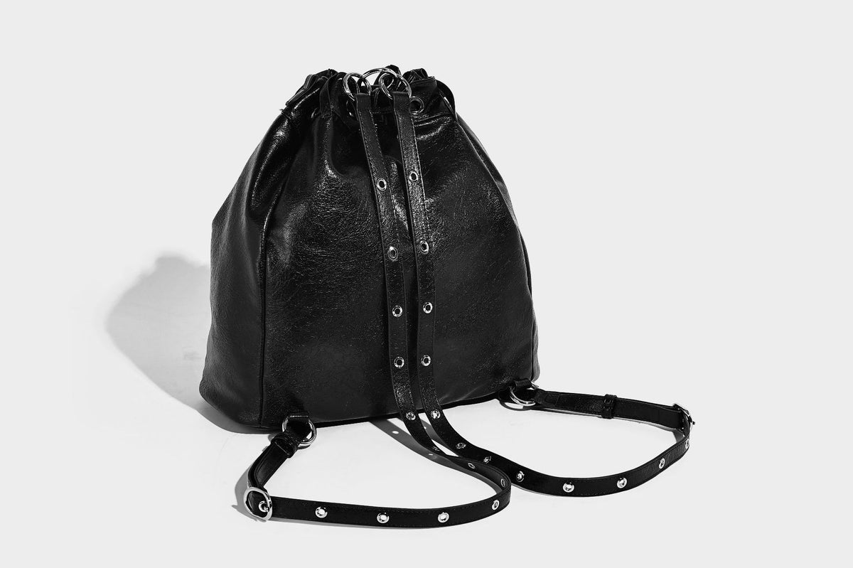 Glossy black convertible bucket backpack with silver grommet-studded straps, drawstring cinch closure, and front zip pocket