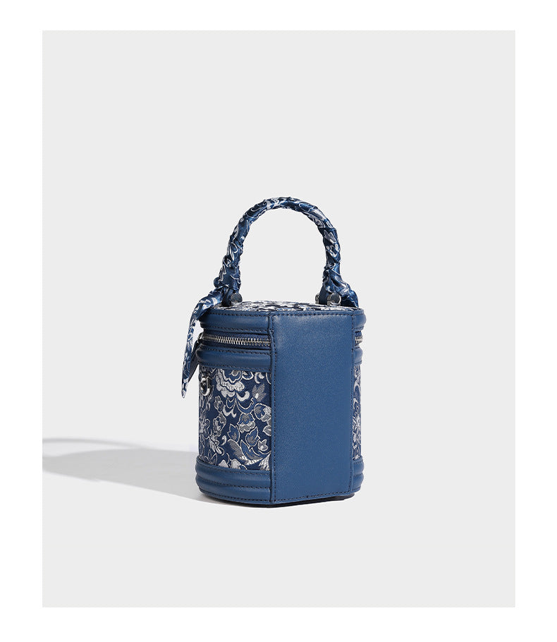 Small cylindrical barrel bag in blue-and-white floral jacquard with leather-look trim, scarf-wrapped top handle, silver-tone