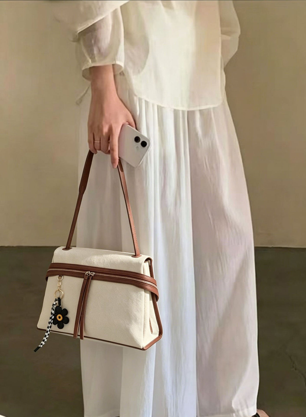 Cream canvas tote bag with brown leather-look trim, dual exterior zip pockets, long shoulder strap, and black flower charm