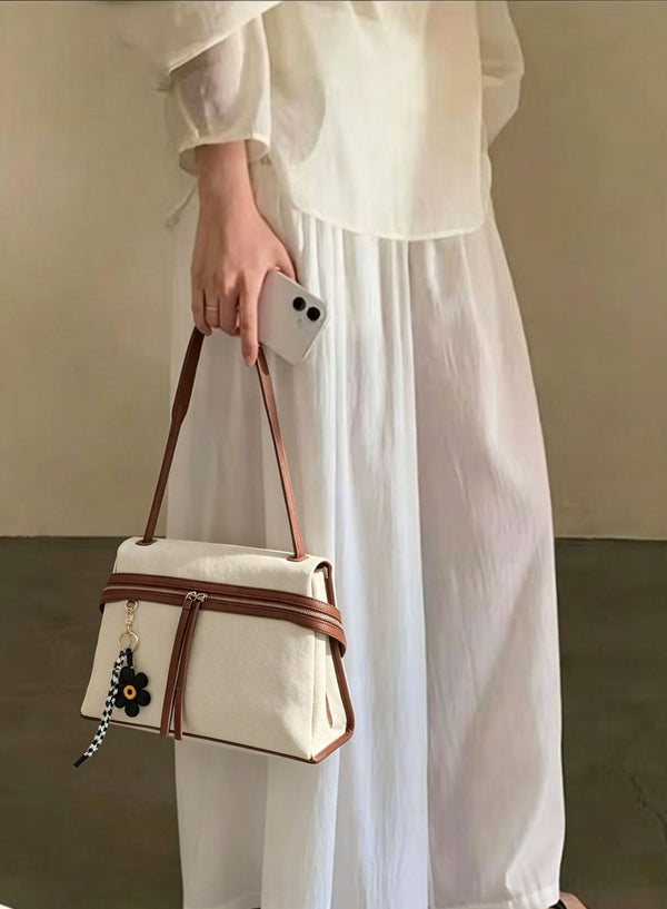 Cream canvas tote bag with brown leather-look trim, dual exterior zip pockets, long shoulder strap, and black flower charm