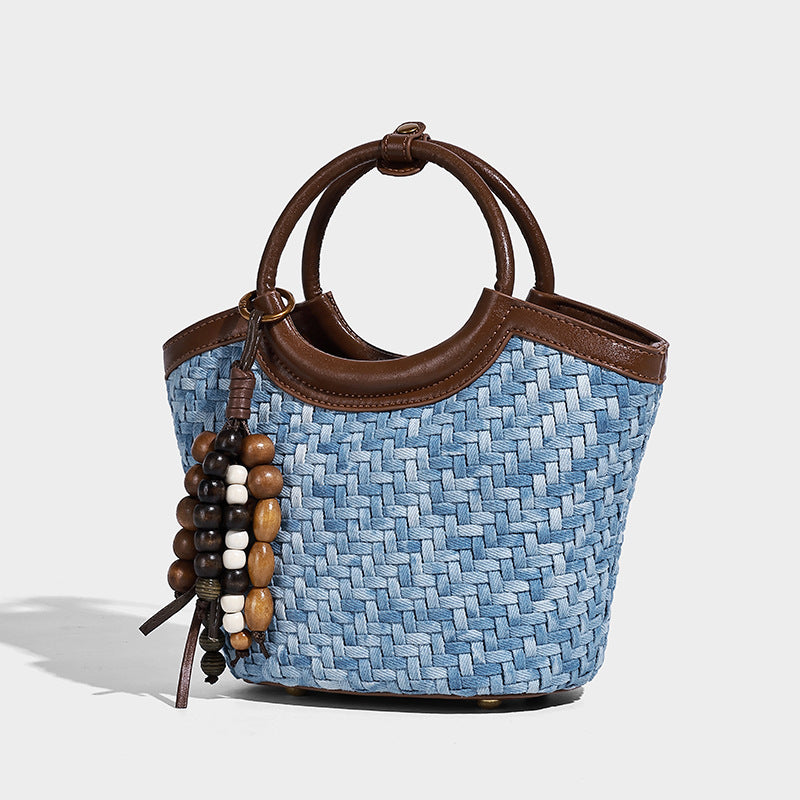 Blue woven denim-look bucket bag with brown leather-look trim, circular top handle, wooden bead tassel charm, and adjustable