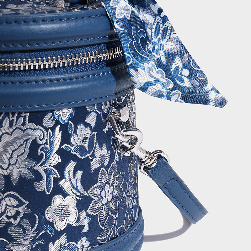 Small cylindrical barrel bag in blue-and-white floral jacquard with leather-look trim, scarf-wrapped top handle, silver-tone