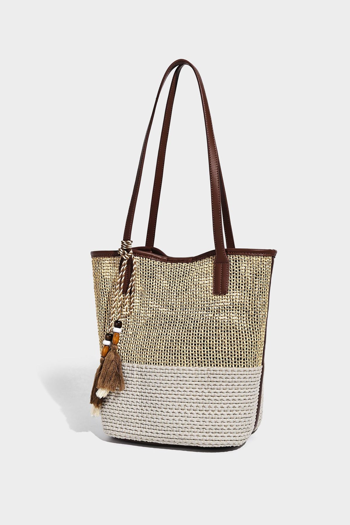 Woven straw-look tote bag in tan and cream with dark brown leather-look trim, long flat shoulder straps, and tassel bead
