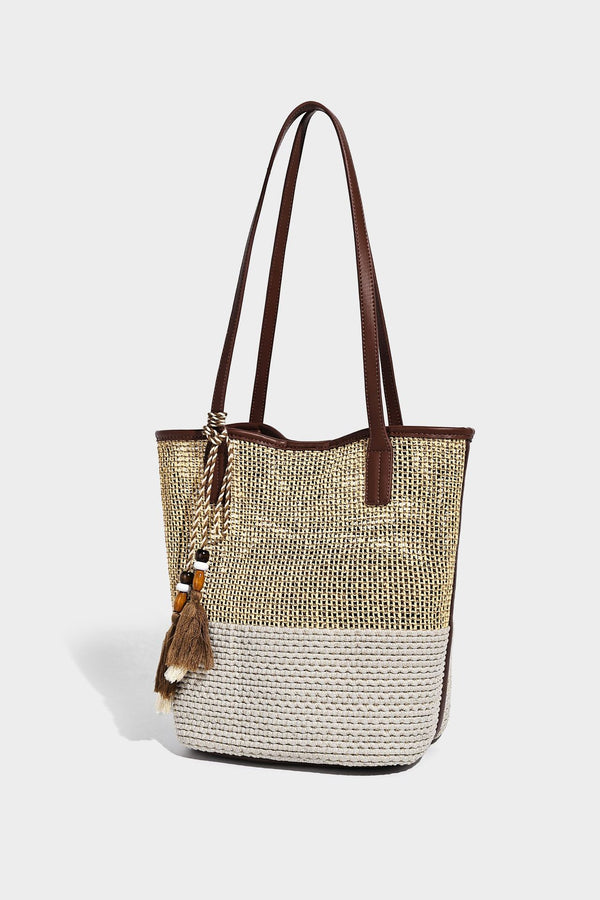 Woven straw-look tote bag in tan and cream with dark brown leather-look trim, long flat shoulder straps, and tassel bead