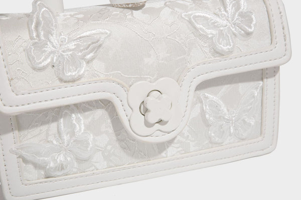 Small structured white flap bag with butterfly embroidery, flower-shaped turn-lock clasp, braided top handle with scarf bow