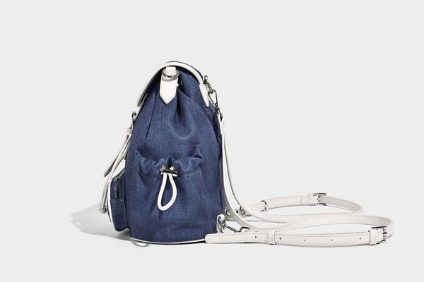Compact denim drawstring backpack with white leather-look piping, flap-top buckle closure, front zip pocket, and silver-tone