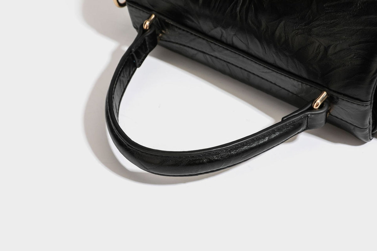 Structured black satchel bag with glossy crinkled leather-look finish, gold-tone emblem flap closure, slim top handle, and
