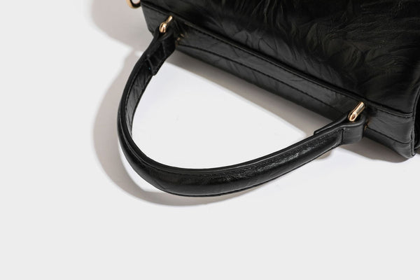 Structured black satchel bag with glossy crinkled leather-look finish, gold-tone emblem flap closure, slim top handle, and