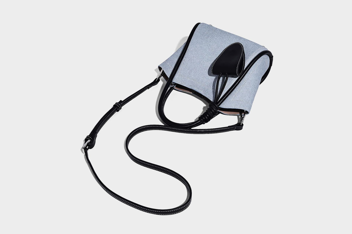 Light blue denim trapezoid tote bag with black leather-look trim, knotted top handles, detachable crossbody strap, front