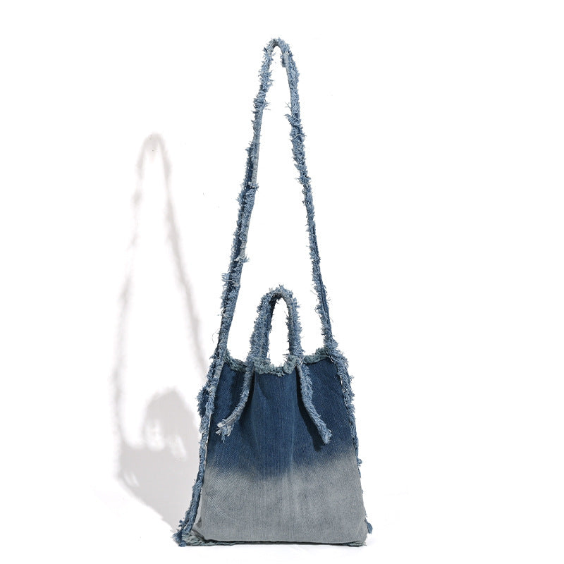 Washed denim fringe crossbody bag with indigo-to-pale-blue gradient and raw fringe trim along edges and shoulder strap