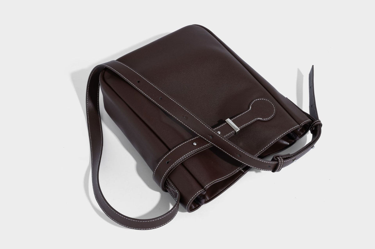 Structured dark brown leather-look bucket bag with white contrast stitching, front wraparound belt buckle strap, and long