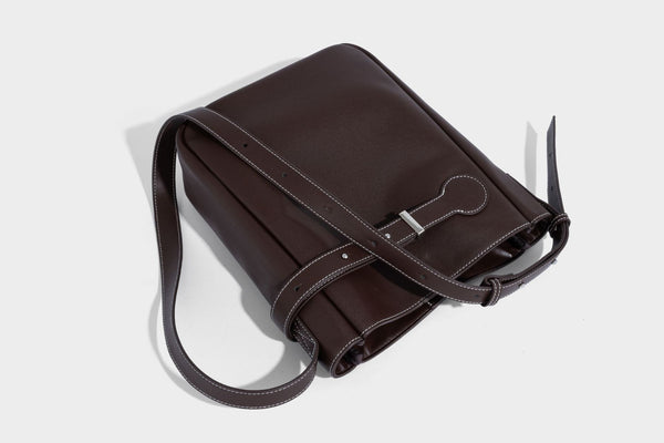 Structured dark brown leather-look bucket bag with white contrast stitching, front wraparound belt buckle strap, and long