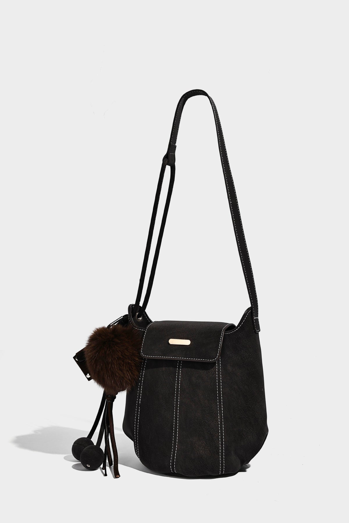 Small matte textured bucket crossbody bag with flap closure, gold-tone bar hardware, contrast stitching, and pompom tassel