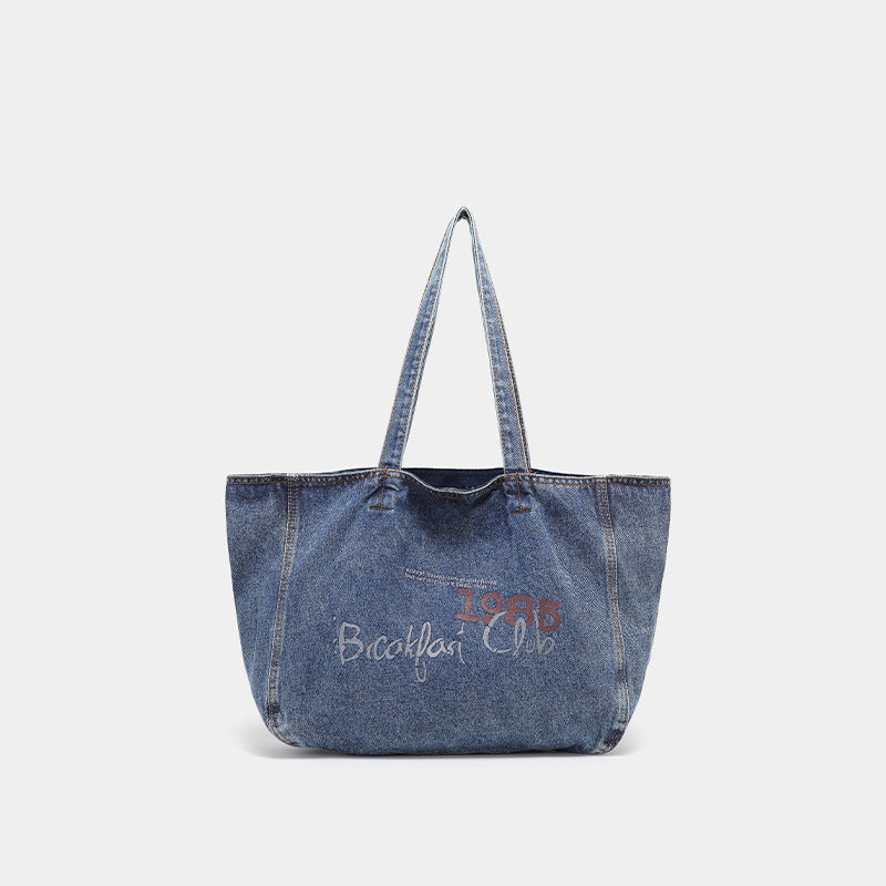 Large washed blue denim tote bag with handwritten-style graphic print, orange contrast stitching, and long fabric shoulder