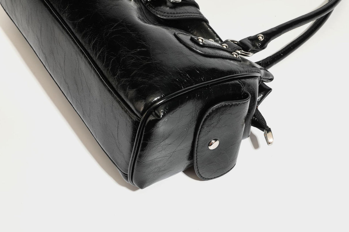 Glänzende, geknautschte Barrel-Satchel mit zwei oberen Tragegriffen, vorderem Klappen-Riemen-Panel, Ring- und Nietenbeschlägen sowie einem abnehmbaren