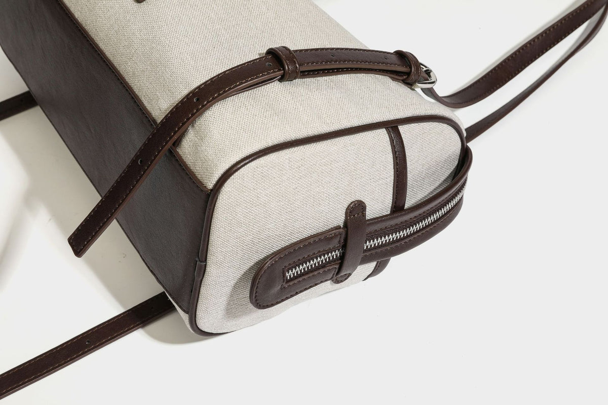 Beige canvas barrel bag with dark brown leather-look trim, silver-tone hardware, top zip closure, and adjustable shoulder