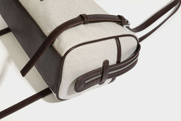 Beige canvas barrel bag with dark brown leather-look trim, silver-tone hardware, top zip closure, and adjustable shoulder