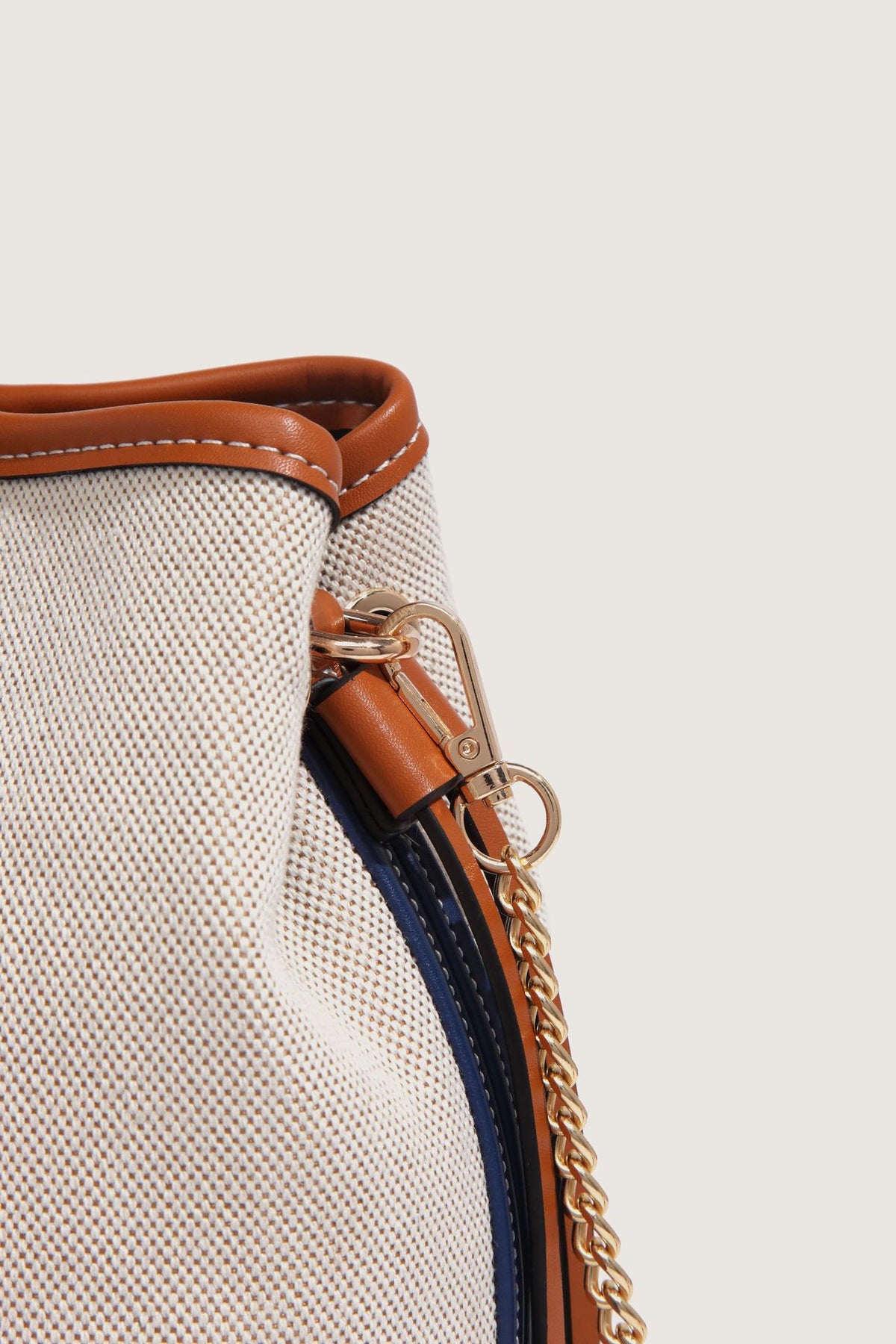 Cream woven-texture canvas bucket bag with navy accent panels, tan leather-look trim, gold-tone chain strap, and tassel