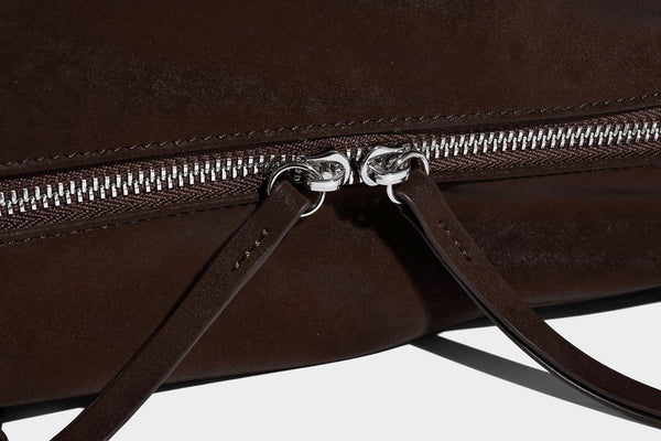 Weiche, zylinderförmige Schultertasche in Dunkelbraun mit silberfarbenen Riemen mit Ösen, Reißverschluss oben, seitlichen Schnallenbeschlägen und Quaste