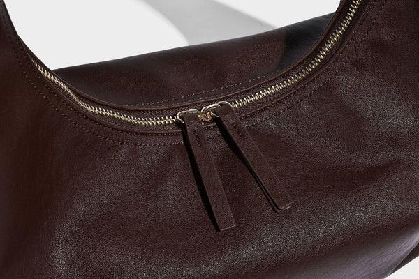 Lässig fallende, sichelförmige Hobo-Schultertasche in glatter dunkelbrauner Lederoptik mit Logo-Anhänger in Goldoptik und verstellbarem Schulterriemen