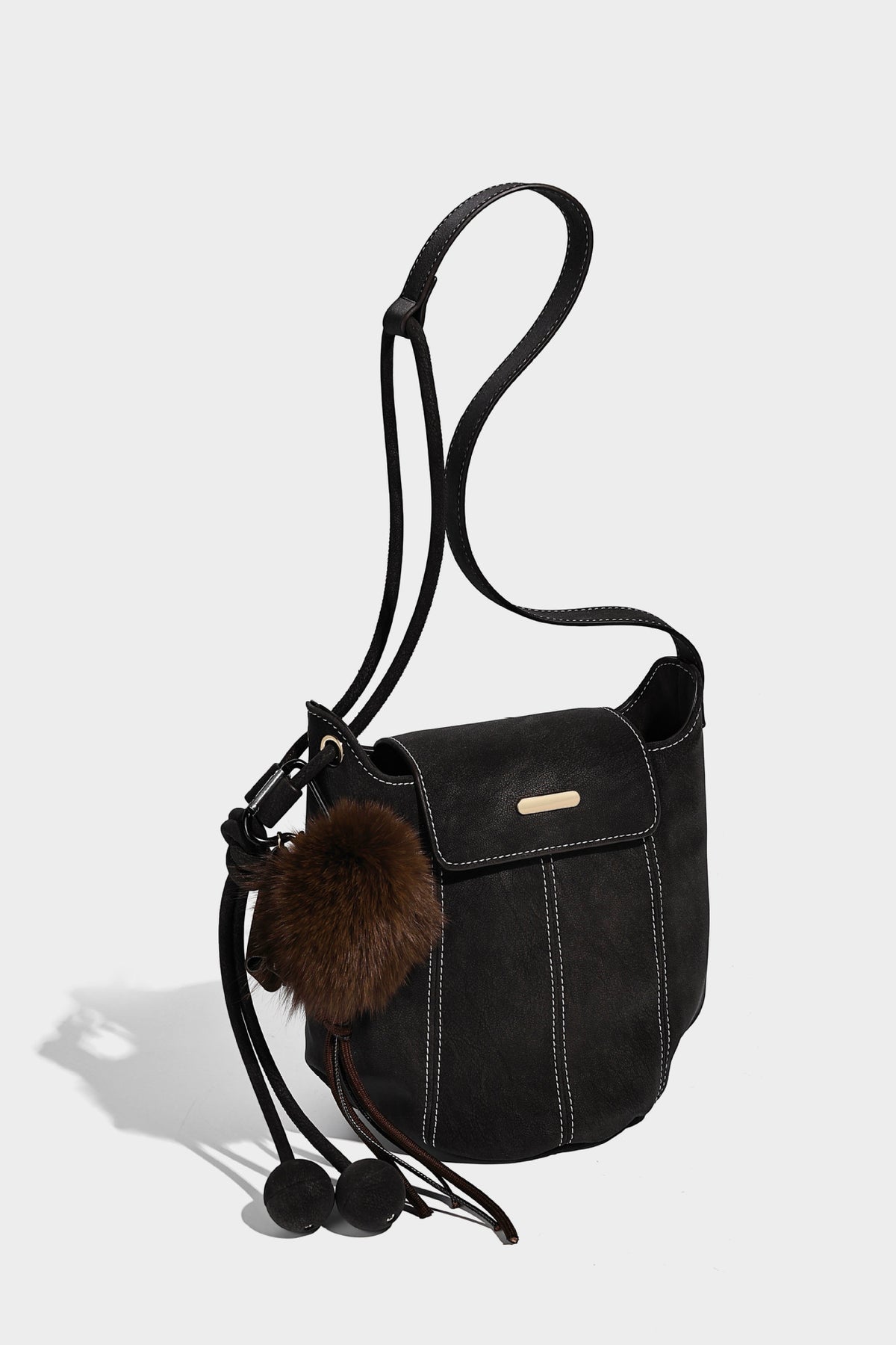 Small matte textured bucket crossbody bag with flap closure, gold-tone bar hardware, contrast stitching, and pompom tassel