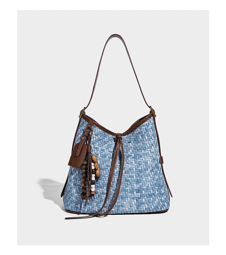 Light blue woven bucket bag with brown leather-look trim, gold-tone hardware, adjustable shoulder strap, and decorative bead