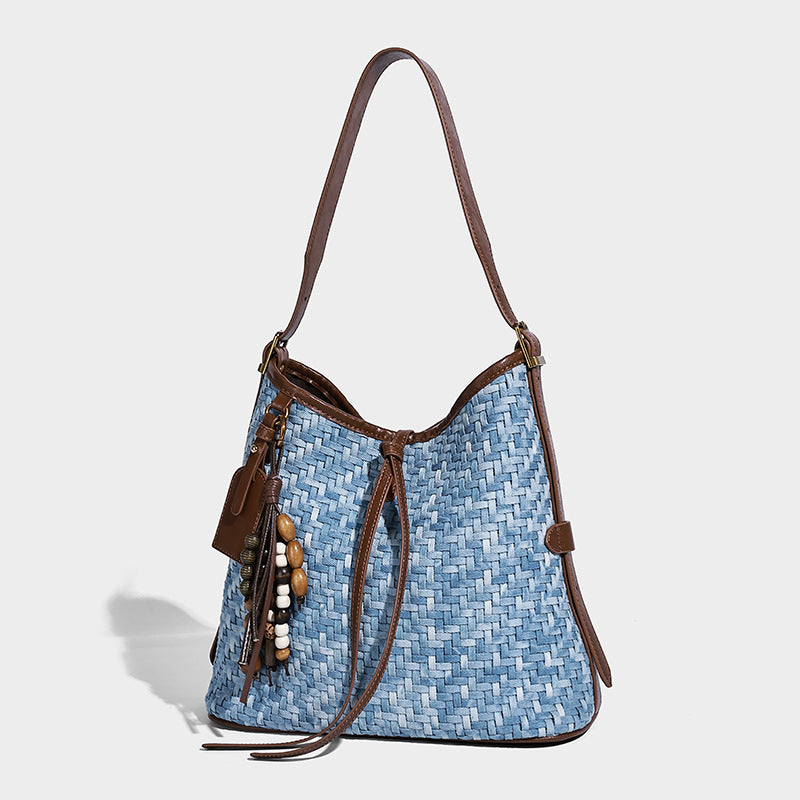 Light blue woven bucket bag with brown leather-look trim, gold-tone hardware, adjustable shoulder strap, and decorative bead
