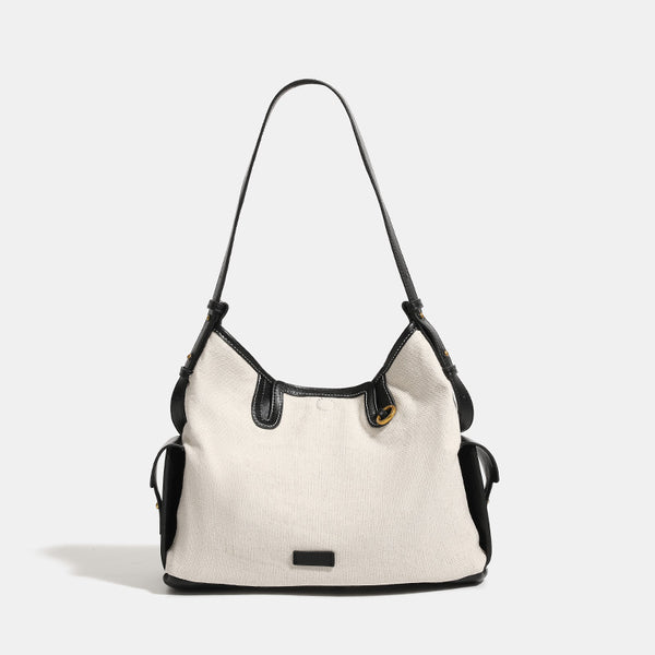 Off-white canvas shoulder bag with black leather-look trim, gold-tone ring hardware, top zip closure, side flap pocket, and
