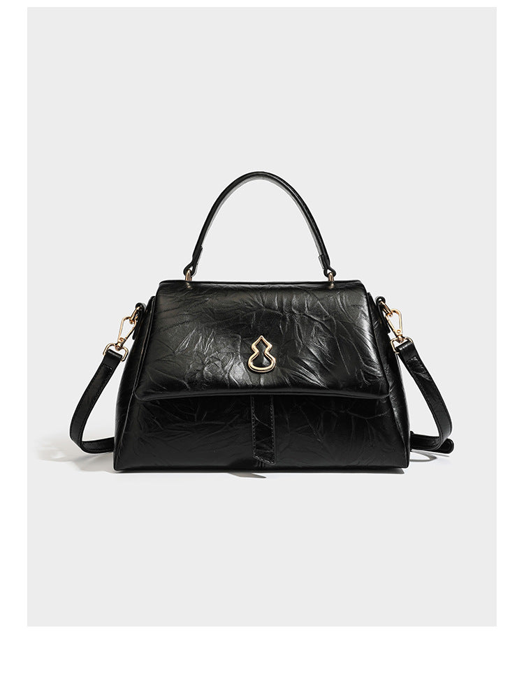 Structured black satchel bag with glossy crinkled leather-look finish, gold-tone emblem flap closure, slim top handle, and