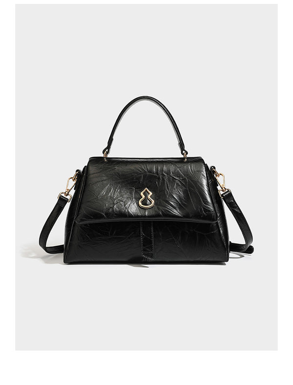 Structured black satchel bag with glossy crinkled leather-look finish, gold-tone emblem flap closure, slim top handle, and