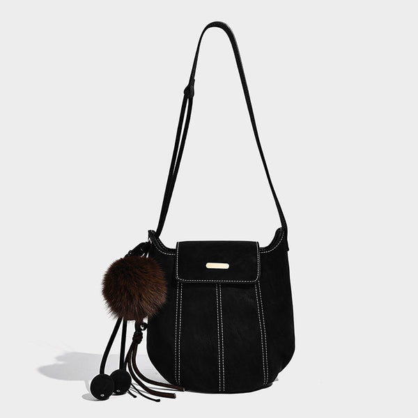 Small matte textured bucket crossbody bag with flap closure, gold-tone bar hardware, contrast stitching, and pompom tassel