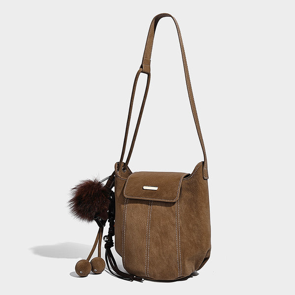 Small matte textured bucket crossbody bag with flap closure, gold-tone bar hardware, contrast stitching, and pompom tassel