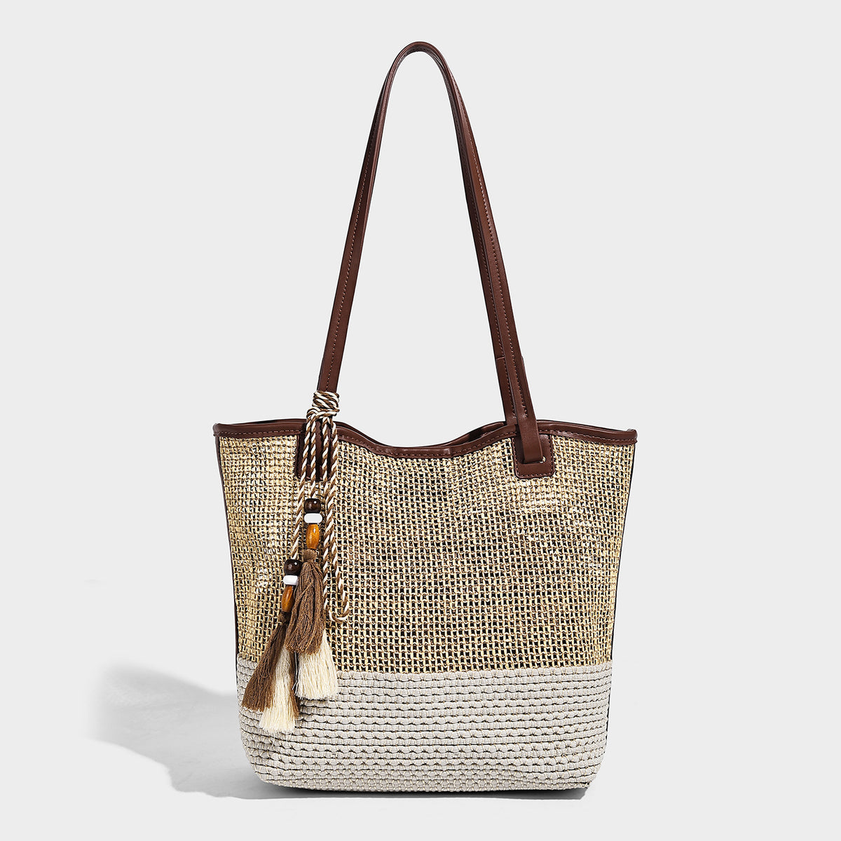 Woven beige and cream tote bag with brown leather-look trim, long double shoulder straps, and knotted tassel charm on front