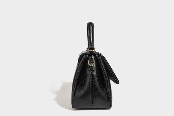 Structured black satchel bag with glossy crinkled leather-look finish, gold-tone emblem flap closure, slim top handle, and