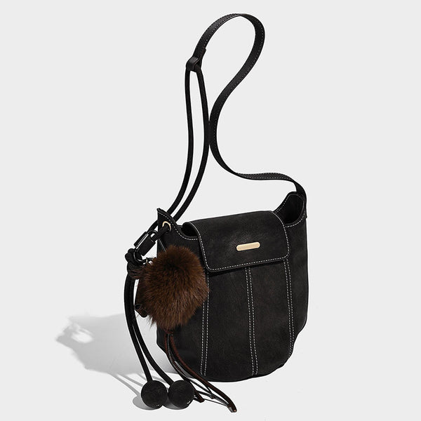 Small matte textured bucket crossbody bag with flap closure, gold-tone bar hardware, contrast stitching, and pompom tassel