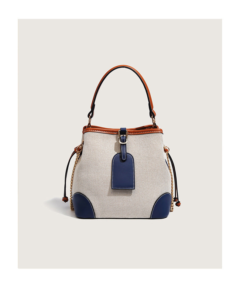 Cream woven-texture canvas bucket bag with navy accent panels, tan leather-look trim, gold-tone chain strap, and tassel
