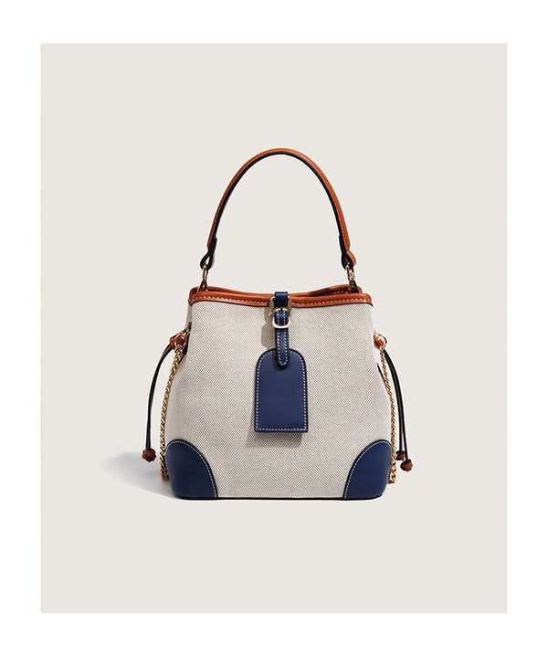 Cream woven-texture canvas bucket bag with navy accent panels, tan leather-look trim, gold-tone chain strap, and tassel