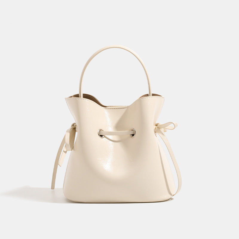 Compact glossy leather-look bucket bag with drawstring bow-tie closure, rounded top handle, and silver-tone grommet hardware