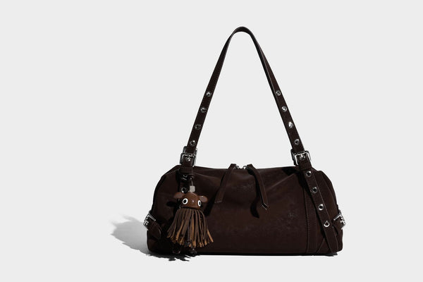 Weiche, zylinderförmige Schultertasche in Dunkelbraun mit silberfarbenen Riemen mit Ösen, Reißverschluss oben, seitlichen Schnallenbeschlägen und Quaste