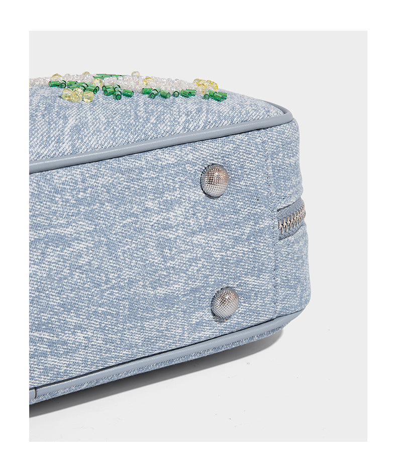 Small structured light blue denim-texture bowling bag with white floral bead embroidery, dual rounded top handles, and