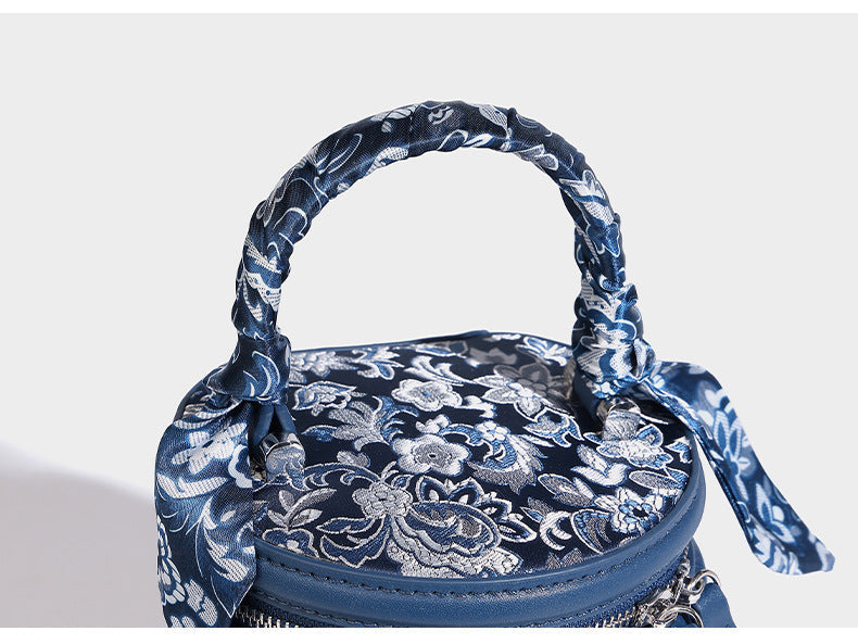Small cylindrical barrel bag in blue-and-white floral jacquard with leather-look trim, scarf-wrapped top handle, silver-tone