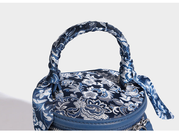 Small cylindrical barrel bag in blue-and-white floral jacquard with leather-look trim, scarf-wrapped top handle, silver-tone